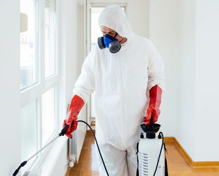 Man in PPE carrying a fluid tank and hose applying pest solution to a white hallway
