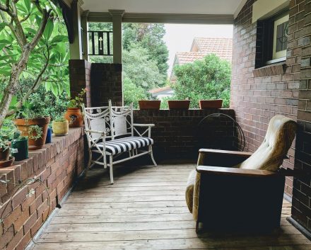 A old brown brick home located in West Ryde with front timber deck will chairs and pot plants